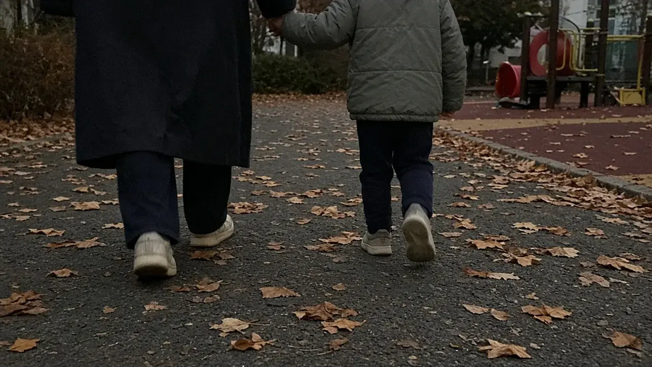 낙엽 깔린 놀이터 옆 길을 어른과 아이가 손을 잡고 걸어가는 뒷모습.